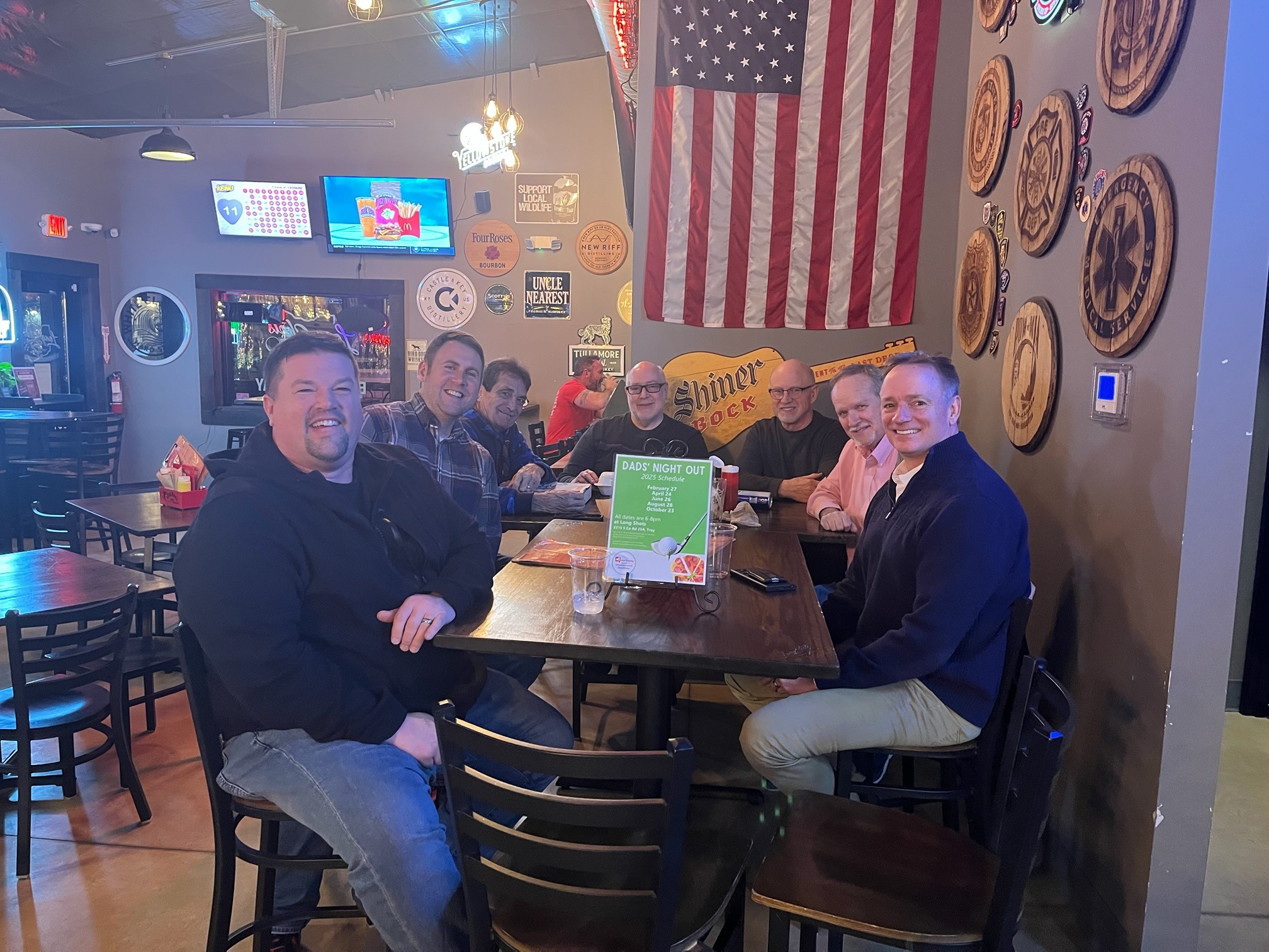 A group of 7 dads sitting at a table in a restaurant smiling at the camera.