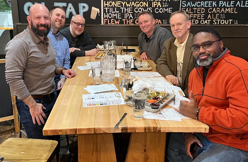 A group of 6 dads sitting at a table in a restaurant smiling at the camera 